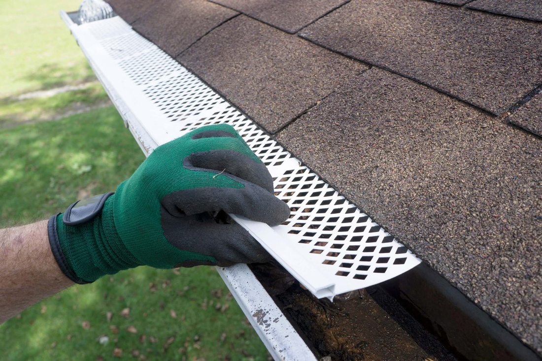 A gloved hand installing a gutter guard on a shingle roof